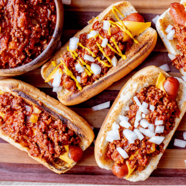 Four chili cheese hot dogs on a wooden cutting board garnished with sliced onions.