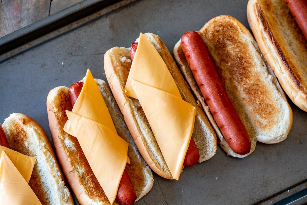Row of smoked hot dogs on toasted hot dog buns topped with two slices of American cheese.