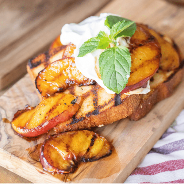 Grilled pound cake topped with grilled peaches, whipped cream, and mint leaves on a wooden cutting board.