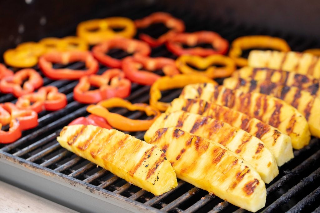 Pineapple spears and bell pepper slices on the grill grates of a grill.