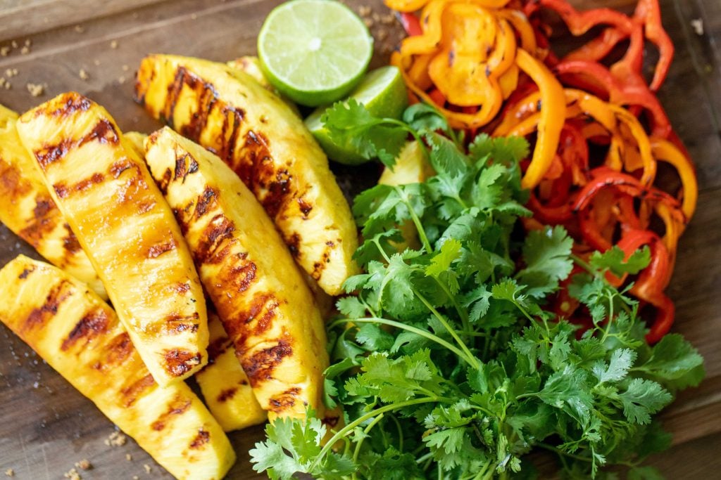 Sliced grilled pineapple and bell peppers next to lime halves and fresh cilantro on a cutting board.