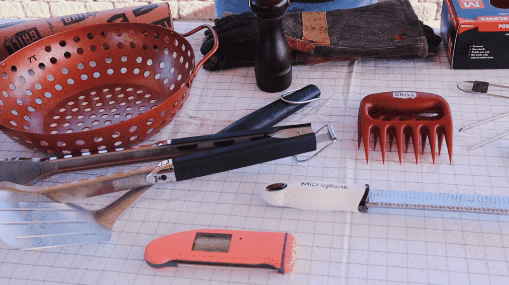 Butcher paper, grill basket, tongs, spatula, meat thermometer, grater, meat claws, and leather gloves on a table.