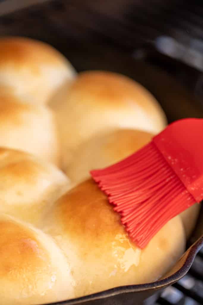 Red basting brush spreading melted butter on golden brown grilled rolls.