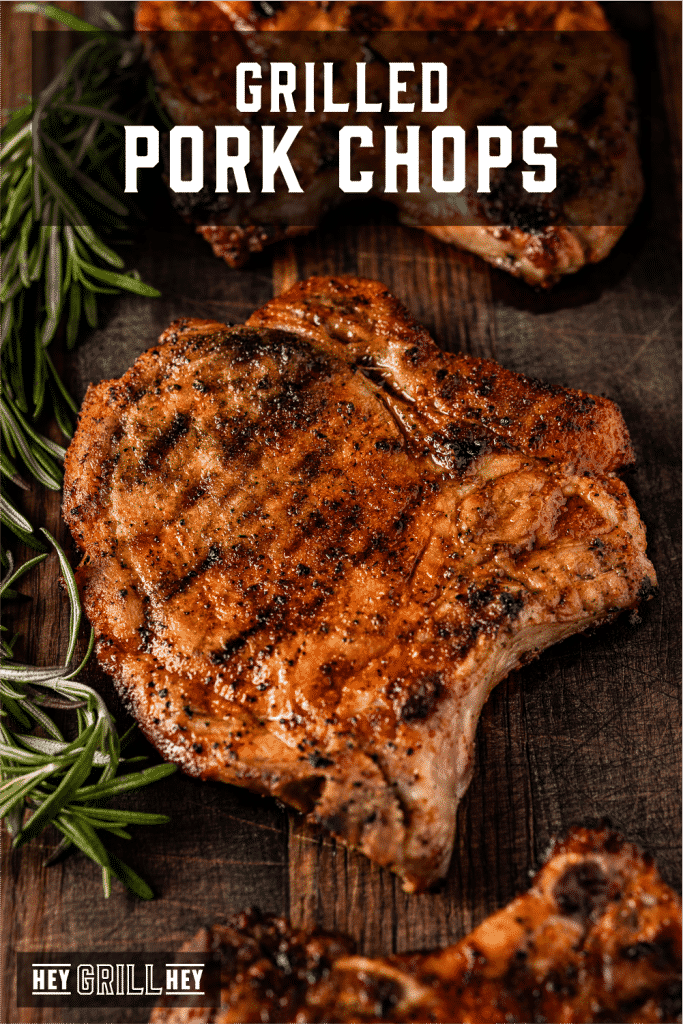 Grilled pork chops on a wooden cutting board surrounded by fresh rosemary with text overlay - Grilled Pork Chops.