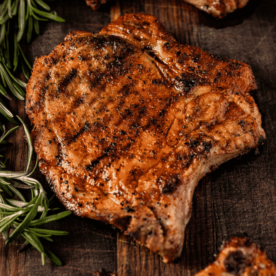 Grilled pork chops on a wooden cutting board surrounded by fresh rosemary.