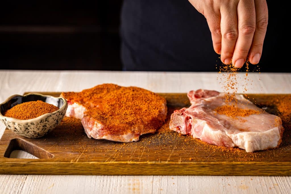 Sweet Rub being sprinkled on pork chops on a wooden cutting board.