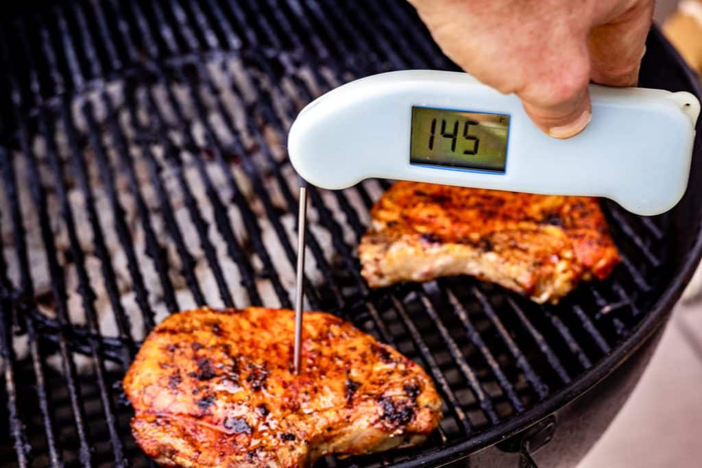 Pork chops on the grill with instant-read thermometer reading temperature of 145 degrees F.