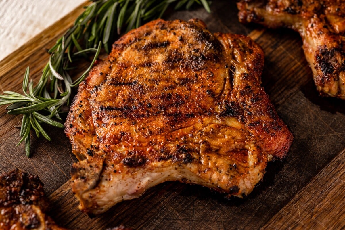 Grilled pork chops on a wooden cutting board surrounded by fresh rosemary.