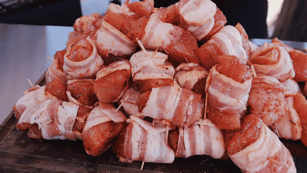 Uncooked bacon wrapped chicken bites secured with toothpicks stacked on a wooden cutting board.