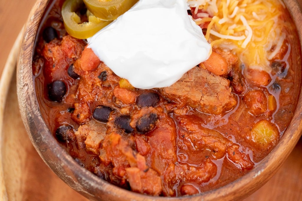 Wooden bowl of brisket chili topped with shredded cheese, sliced peppers, and a dollop of sour cream.