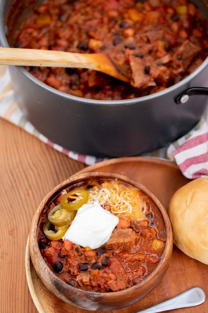 Bowl of brisket chili on a plate next to a roll and metal spoon with a pot of brisket chili in the background.