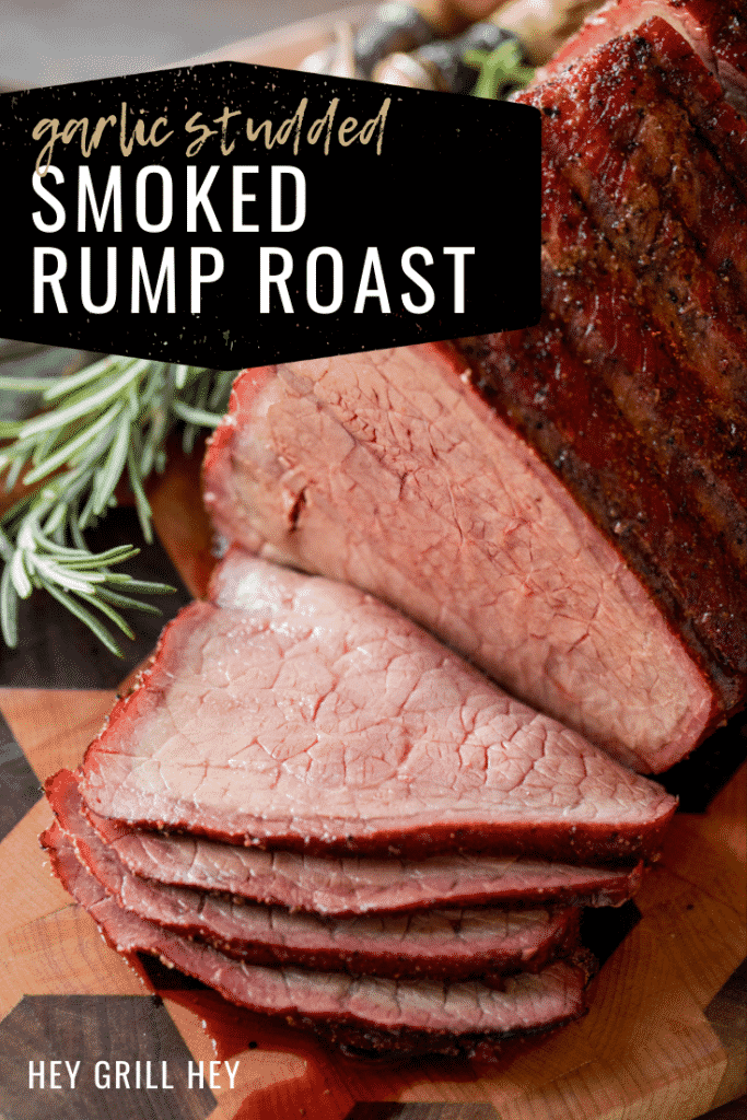 Smoked and sliced beef rump roast on a wooden cutting board next to fresh herbs and metal utensils. Text overlay reads: Garlic Studded Smoked Rump Roast.