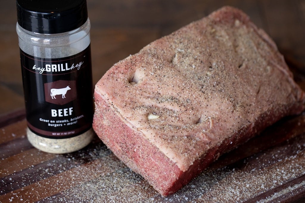 Whole uncooked rump roast inserted with garlic cloves and seasoned with Hey Grill Hey Beef Seasoning next to a bottle of Beef Seasoning.
