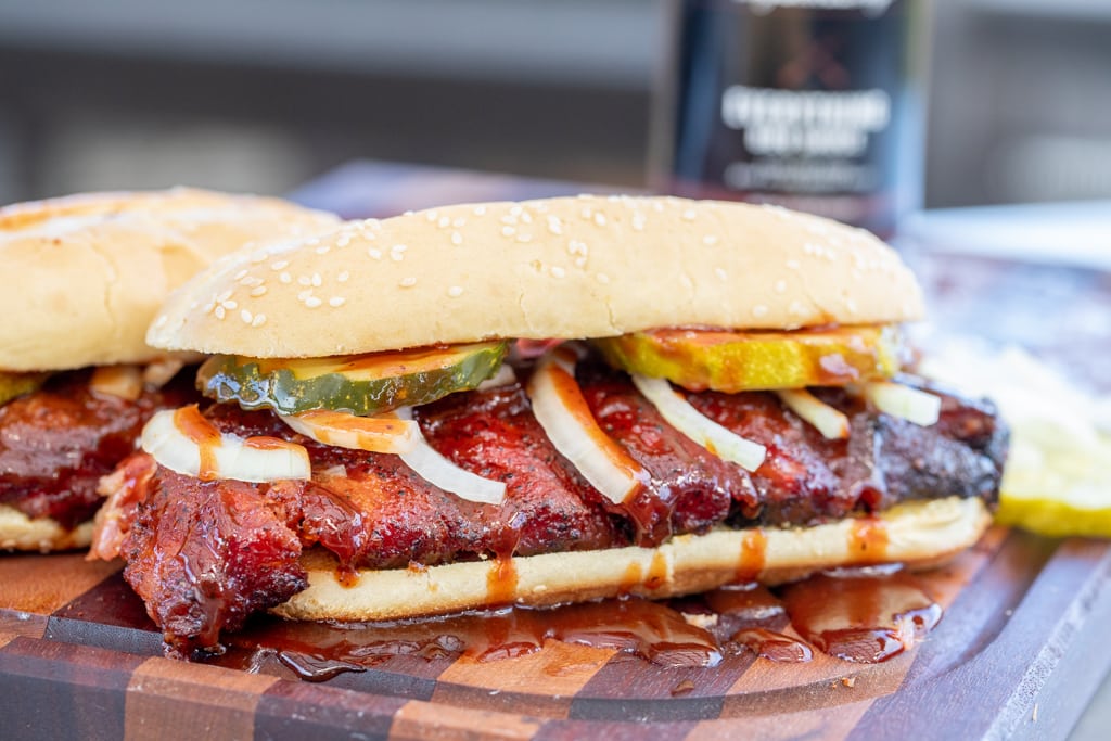 Smoked rib sandwich topped with BBQ sauce and sliced onions and pickles on a wooden cutting board.