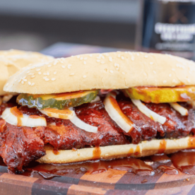 Smoked rib sandwich topped with BBQ sauce and sliced onions and pickles on a wooden cutting board.