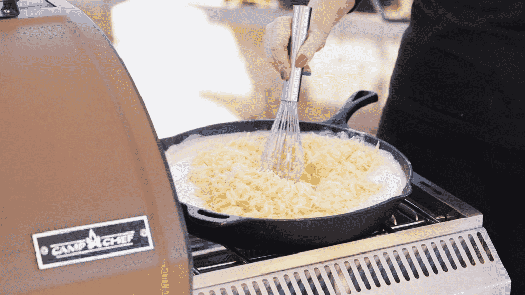 Shredded white cheddar and gouda cheese being whisked into milk and roux in a cast iron skillet.