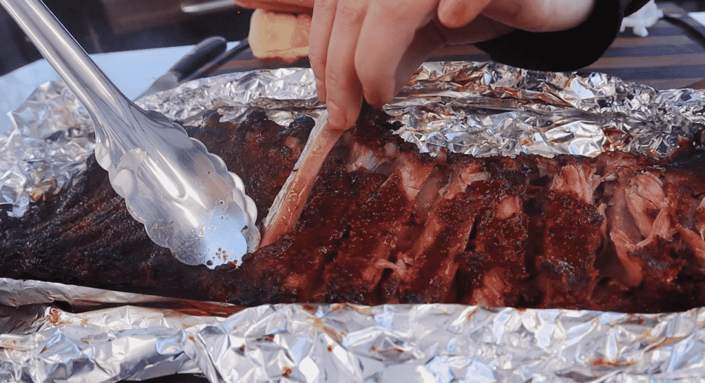 Metal tongs pulling a rib bone out of a whole rack of ribs.