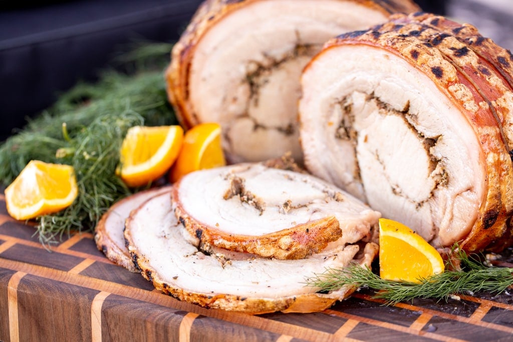 Sliced porchetta surrounded by lime wedges and fresh herbs on a wooden cutting board.