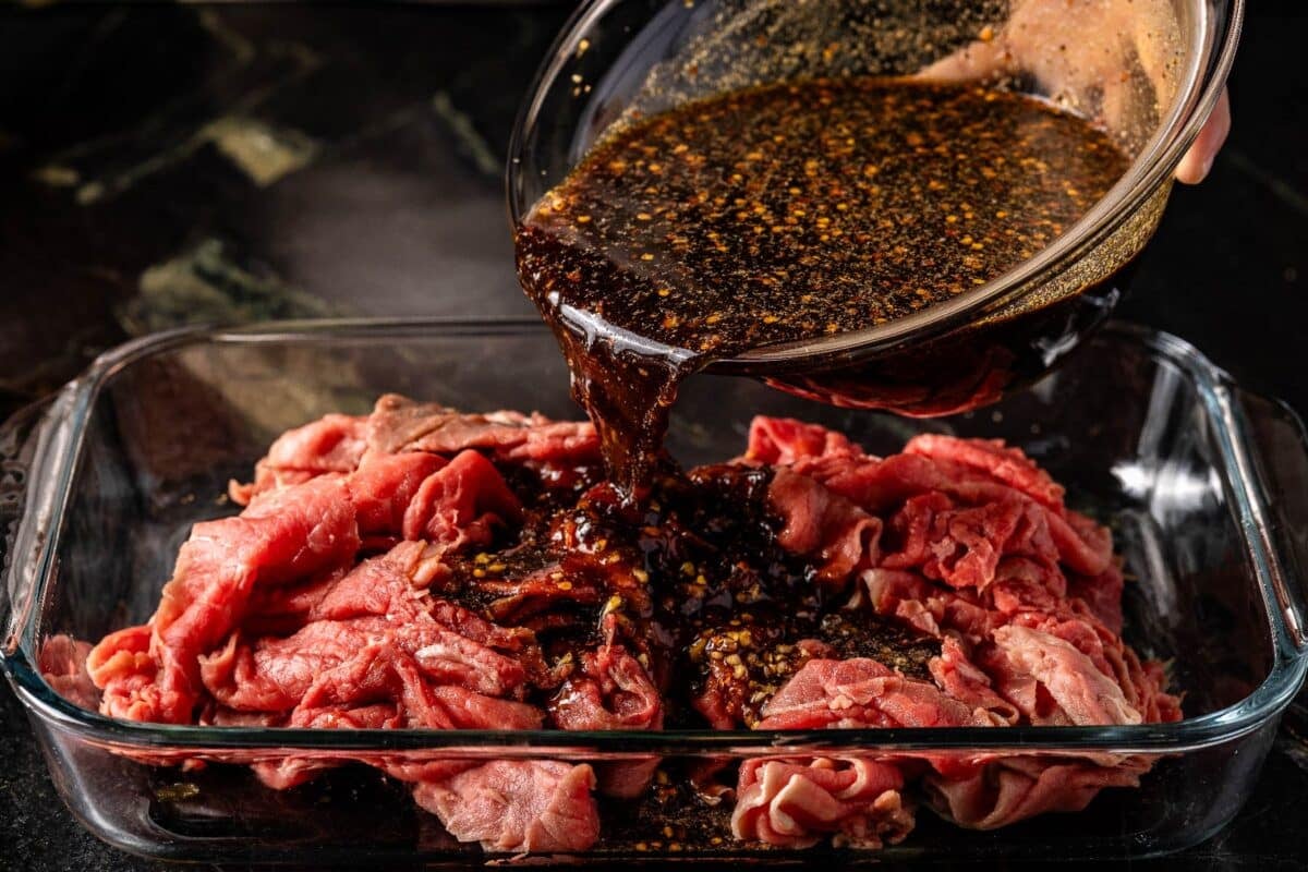 Beef slices in a glass baking dish with a marinade being poured on them from a bowl.