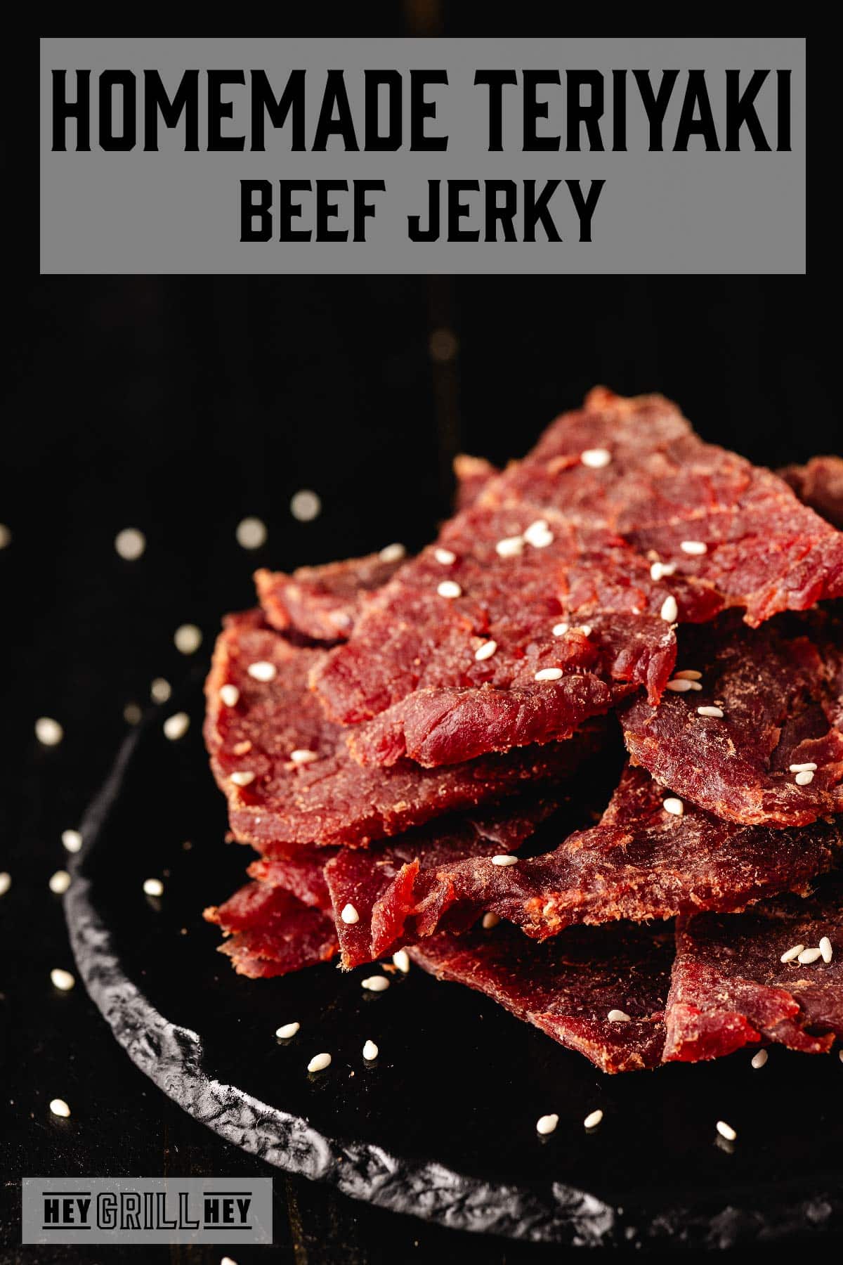 Jerky slices piled on a black serving platter, sprinkled with salt flakes. The text overlay reads "Homemade Teriyaki Beef Jerky" at the top, and "Hey Grill Hey" at the bottom.