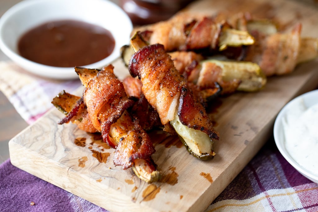 Bacon wrapped pickles stacked on a wooden board with a small bowl of BBQ sauce in the background.