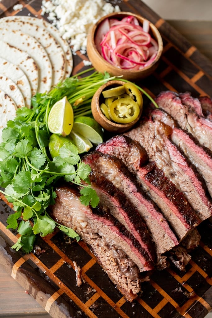 Slices of beef brisket, fresh cilantro, flour tortillas, sliced jalapenos, and a small bowl of pickled red onions arranged on a large wooden cutting board.