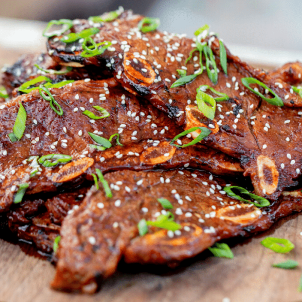 Kalbi garnished with sliced green onions and sesame seeds stacked on a round wooden serving board.