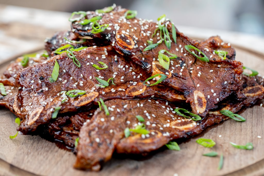 Kalbi garnished with sliced green onions and sesame seeds stacked on a round wooden serving board.