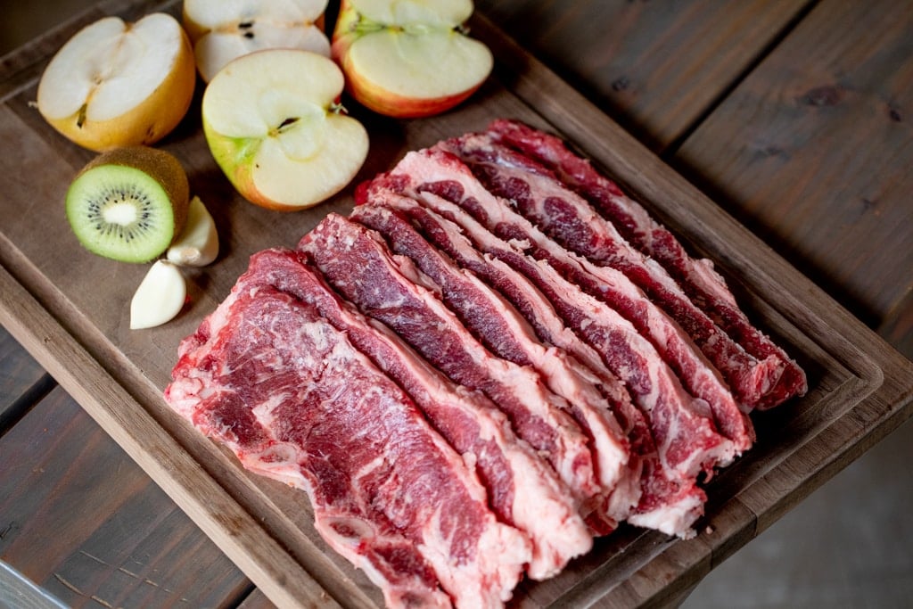 Sliced short ribs lined up on a rectangular wooden cutting board next to four apple halves and sliced kiwi.