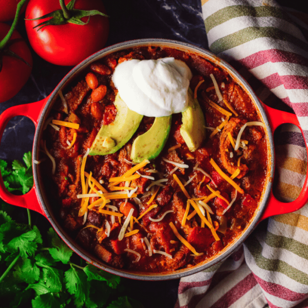 Overhead shot of smoked tri tip chili in a red dutch oven topped with shredded cheese, sliced avocado and a dollop of sour cream.