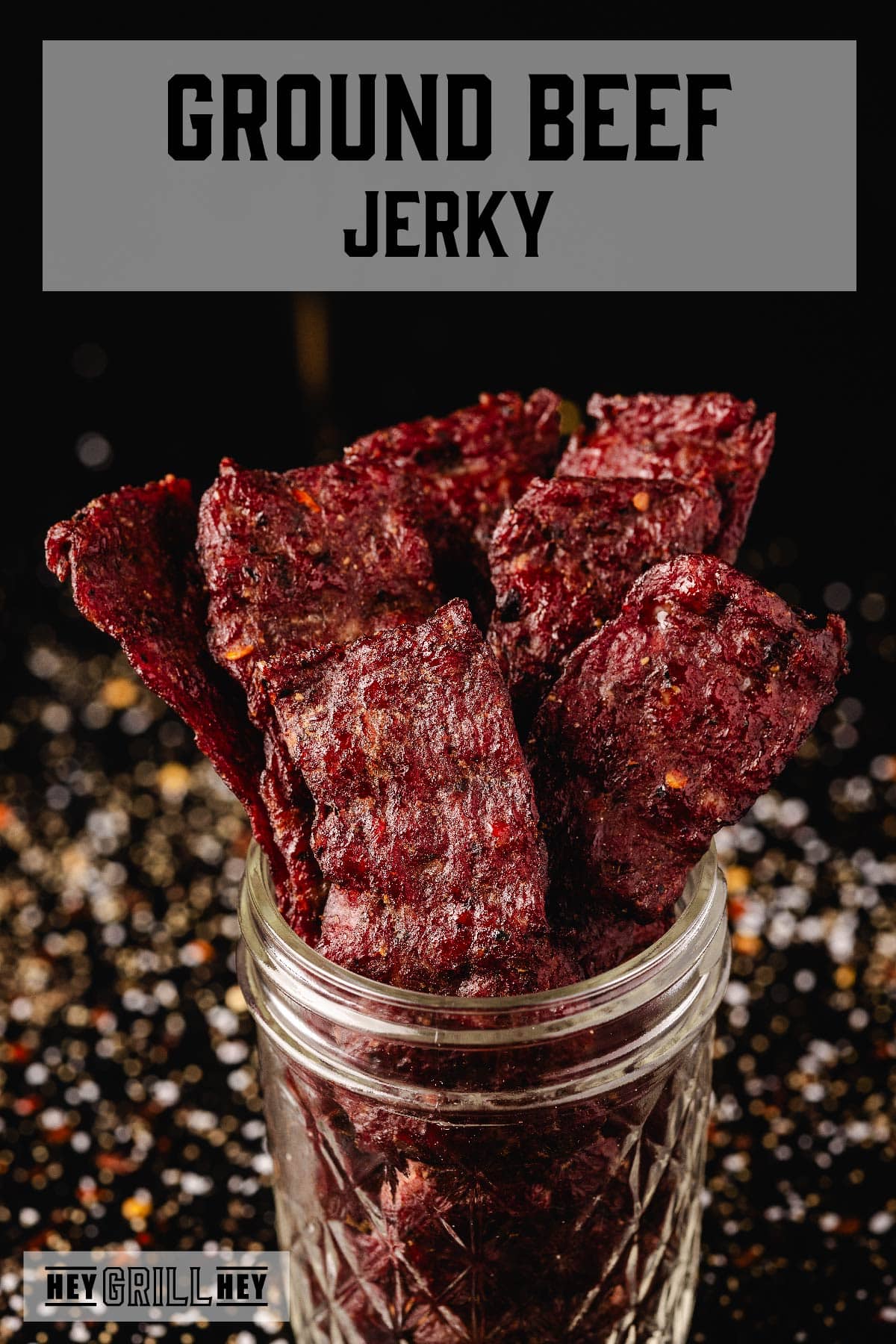 Sticks of jerky in a glass jar on a black surface sprinkled with seasonings. The text overlay reads "Ground Beef Jerky" at the top, and "Hey Grill Hey" at the bottom.