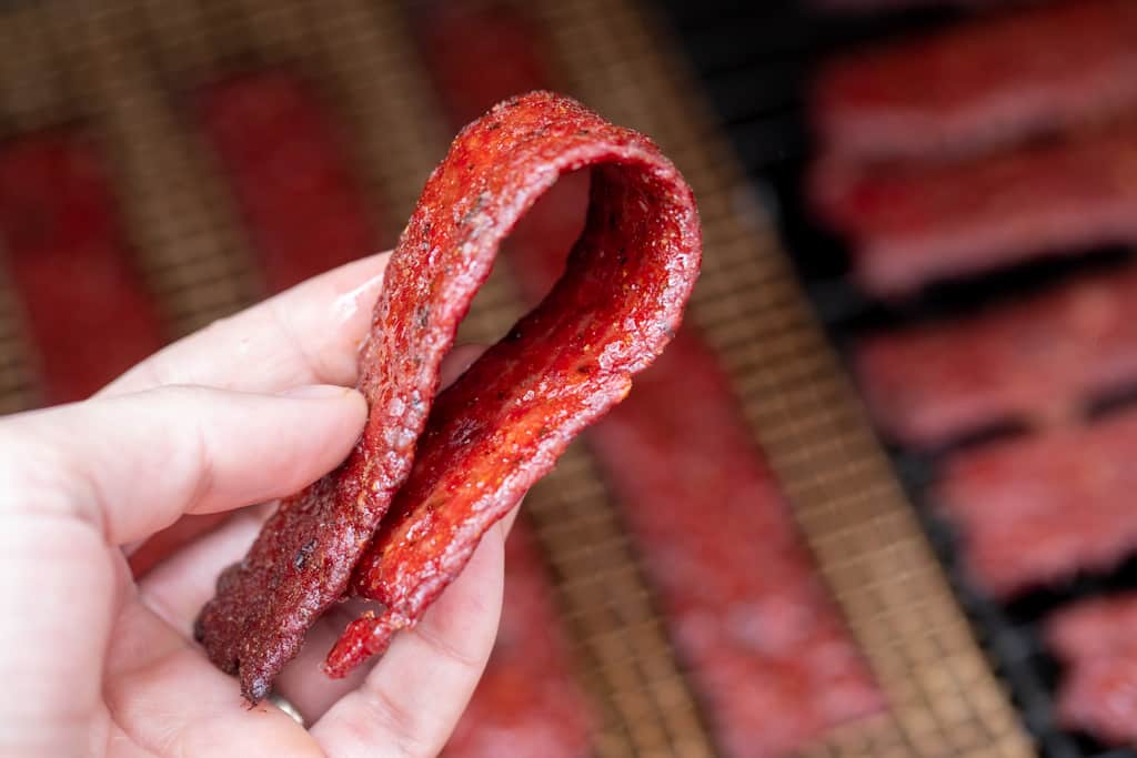 Once slice of ground beef jerky being folded in half with a rack of ground beef jerky strips in the background.
