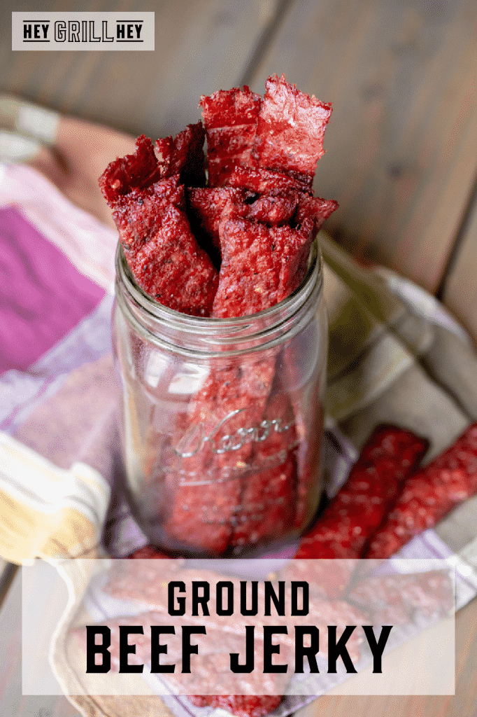 Strips of ground beef jerky in a glass mason jar on a colorful kitchen towel. Text overlay reads: Ground Beef Jerky.