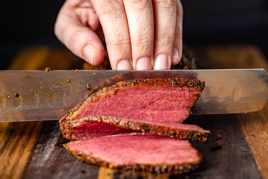 Chef knife slicing through pastrami on a wooden cutting board.