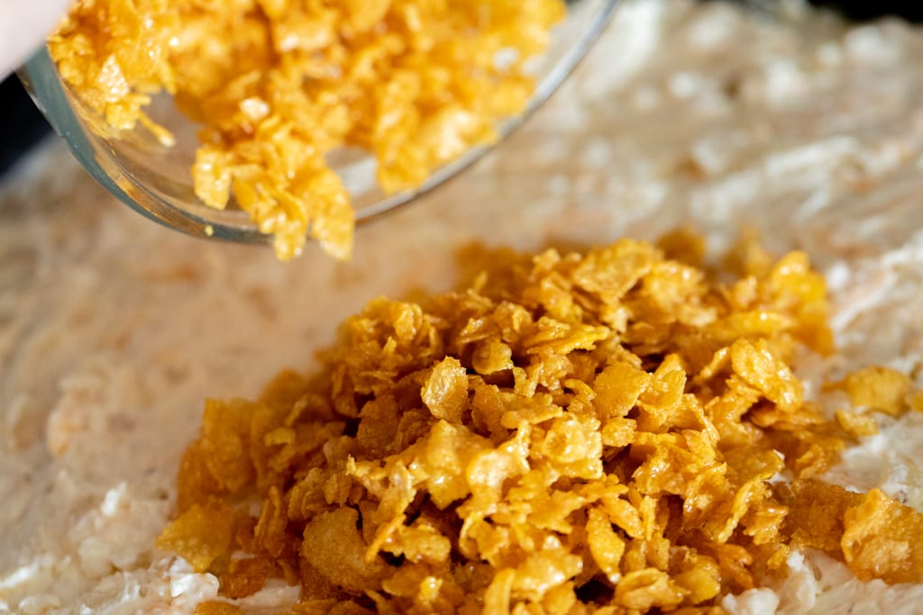 Corn flakes being poured on top of a potato and cheese mixture.