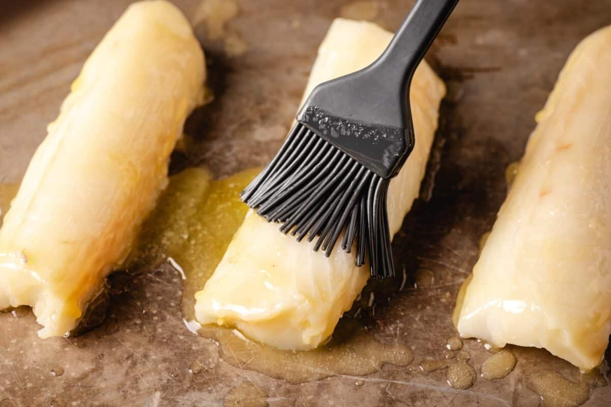 Three raw fish fillets on a cutting board, with a basting brush rubbing butter on the middle one.