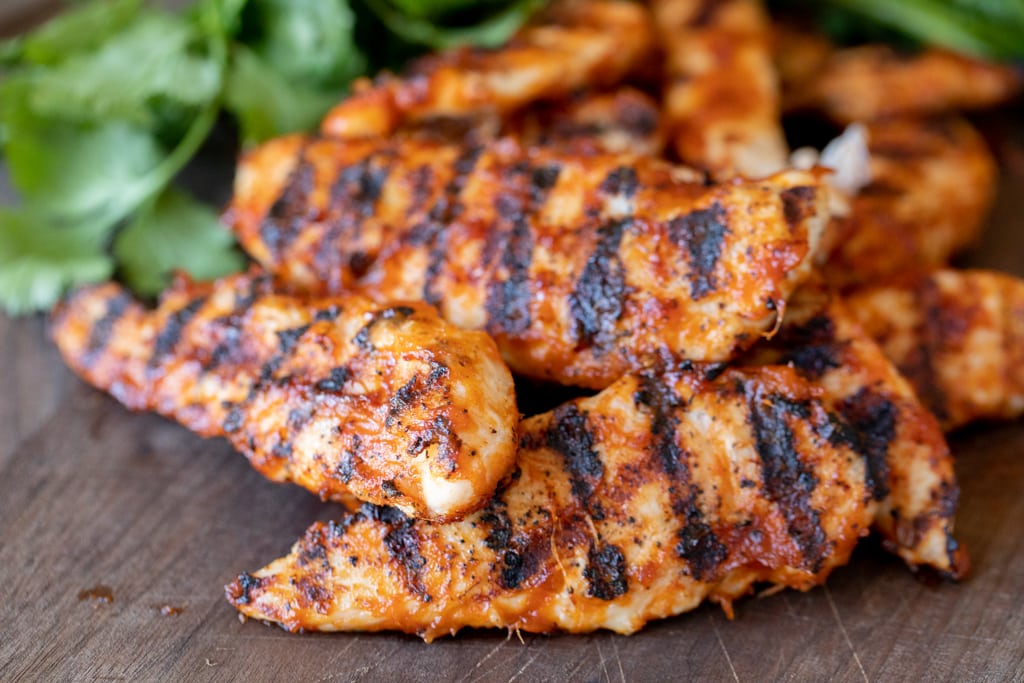 Grilled chicken tenders stacked on a wooden cutting board with fresh herbs in the background.