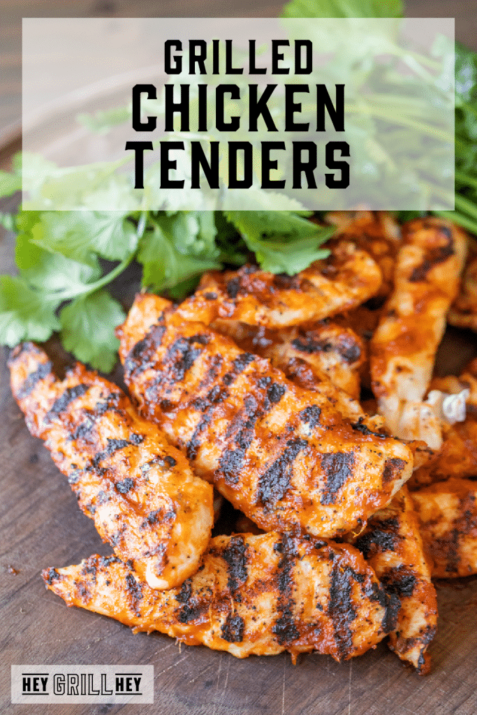Grilled chicken tenders stacked on a wooden cutting board with fresh herbs in the background with text overlay - Grilled Chicken Tenders.
