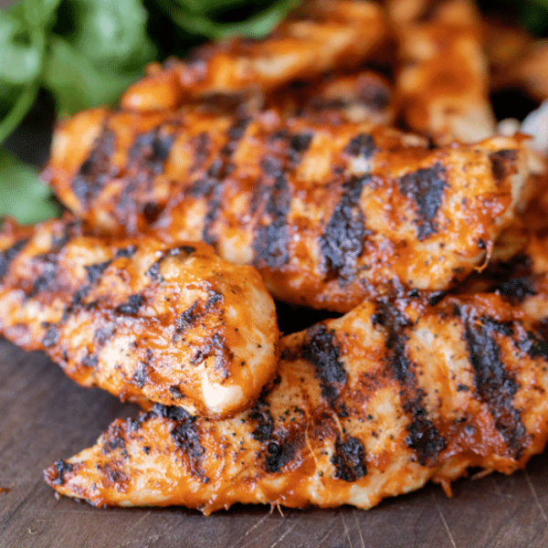 Grilled chicken tenders stacked on a wooden cutting board with fresh herbs in the background.