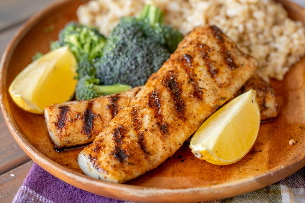 Two grilled mahi mahi filets on a wood plate surrounded by broccoli, rice, and lemon wedges.