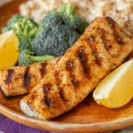Two grilled mahi mahi filets on a plate surrounded by broccoli, rice, and lemon wedges.