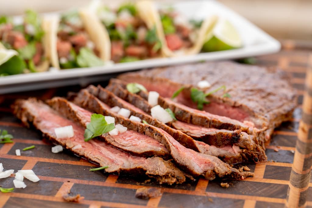 Sliced flank steak on a wooden cutting board with flank steak tacos in the background.