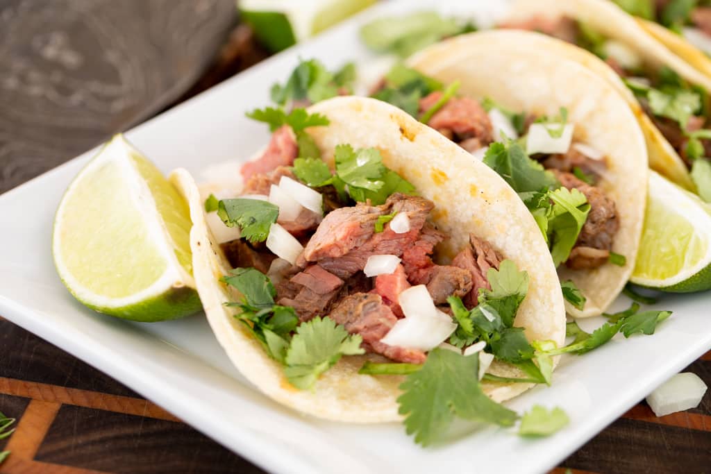 Two flank steak tacos topped with sliced onions and fresh cilantro on a white plate.