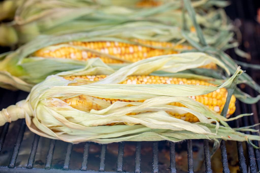 Seasoned corn in the husks on the grill grates of a smoker.