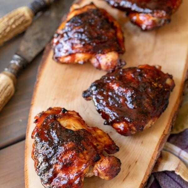 Four BBQ chicken thighs on a long, wooden cutting board.