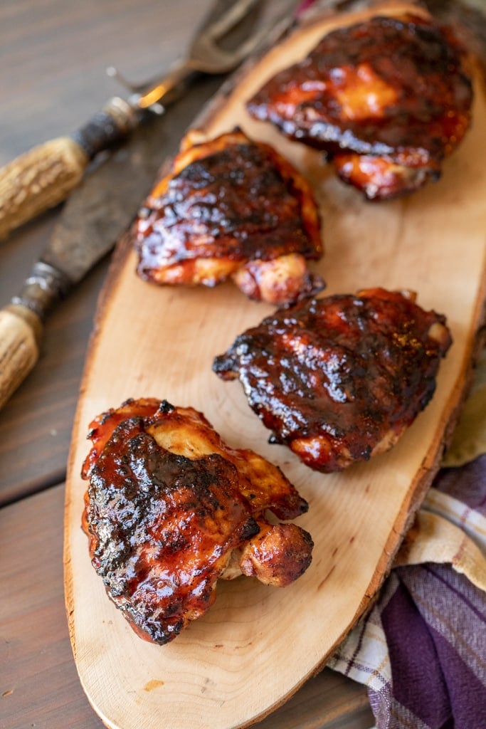 Four BBQ chicken thighs on a long, wooden cutting board.
