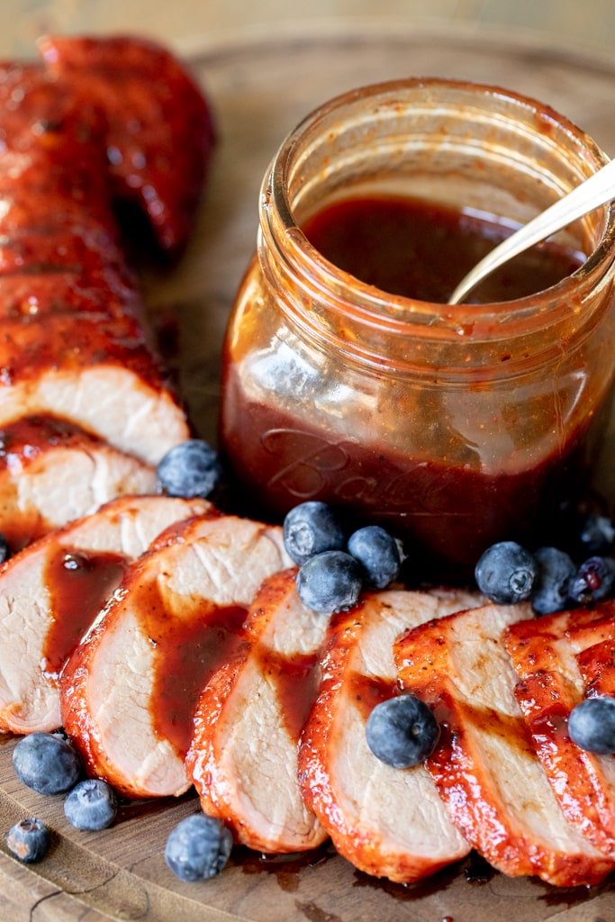 Blueberry BBQ Sauce drizzled over a sliced pork tenderloin with a glass mason jar of blueberry BBQ sauce in the background.