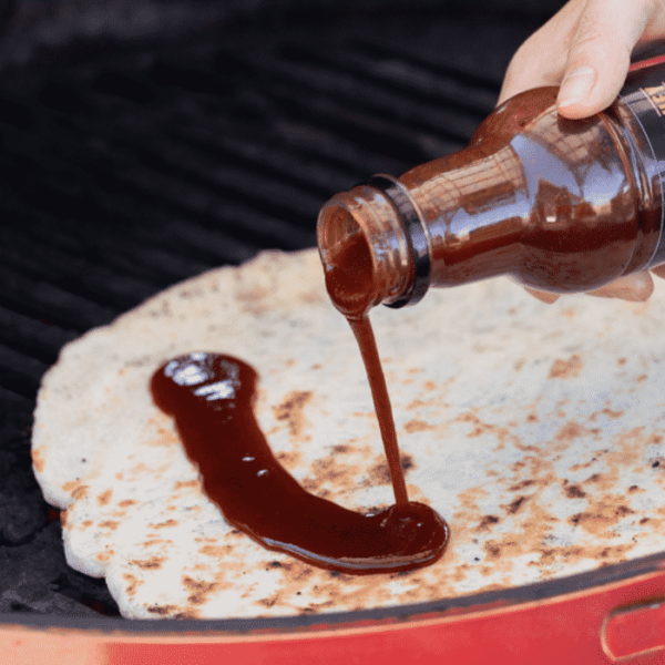 Pizza dough on a gas grill being drizzled with BBQ sauce.