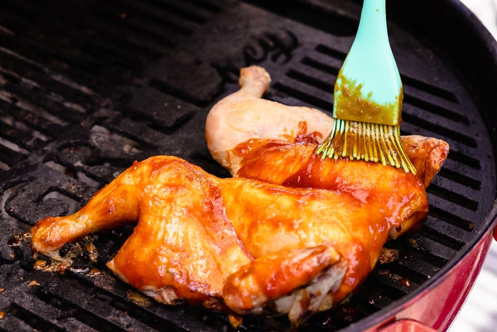 Spatchcocked chicken on a charcoal grill being basted with BBQ sauce.