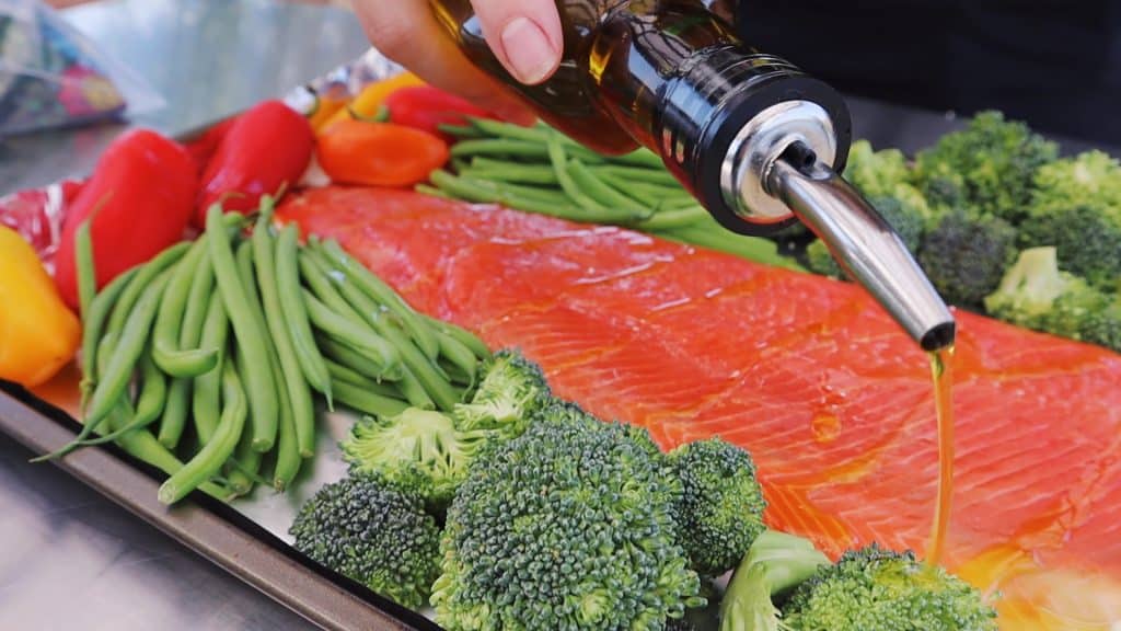 Olive oil being drizzled on a salmon fillet, broccoli, green beans, and peppers on a sheet pan.